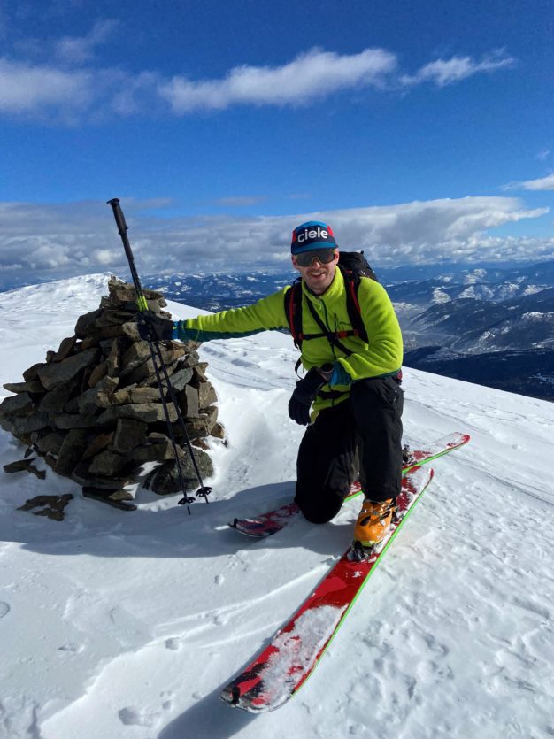 Alexander Spraggs crouched on skis at the top of a snowy mountain next to a pile of stones on a bluebird day.