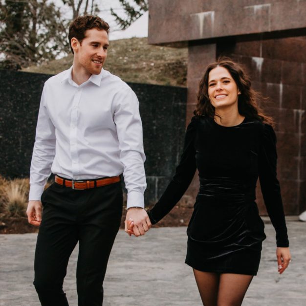 Megan Santano in a black dress holding hands with her partner, a tall white man with brown hair in a white button up shirt and black pants.