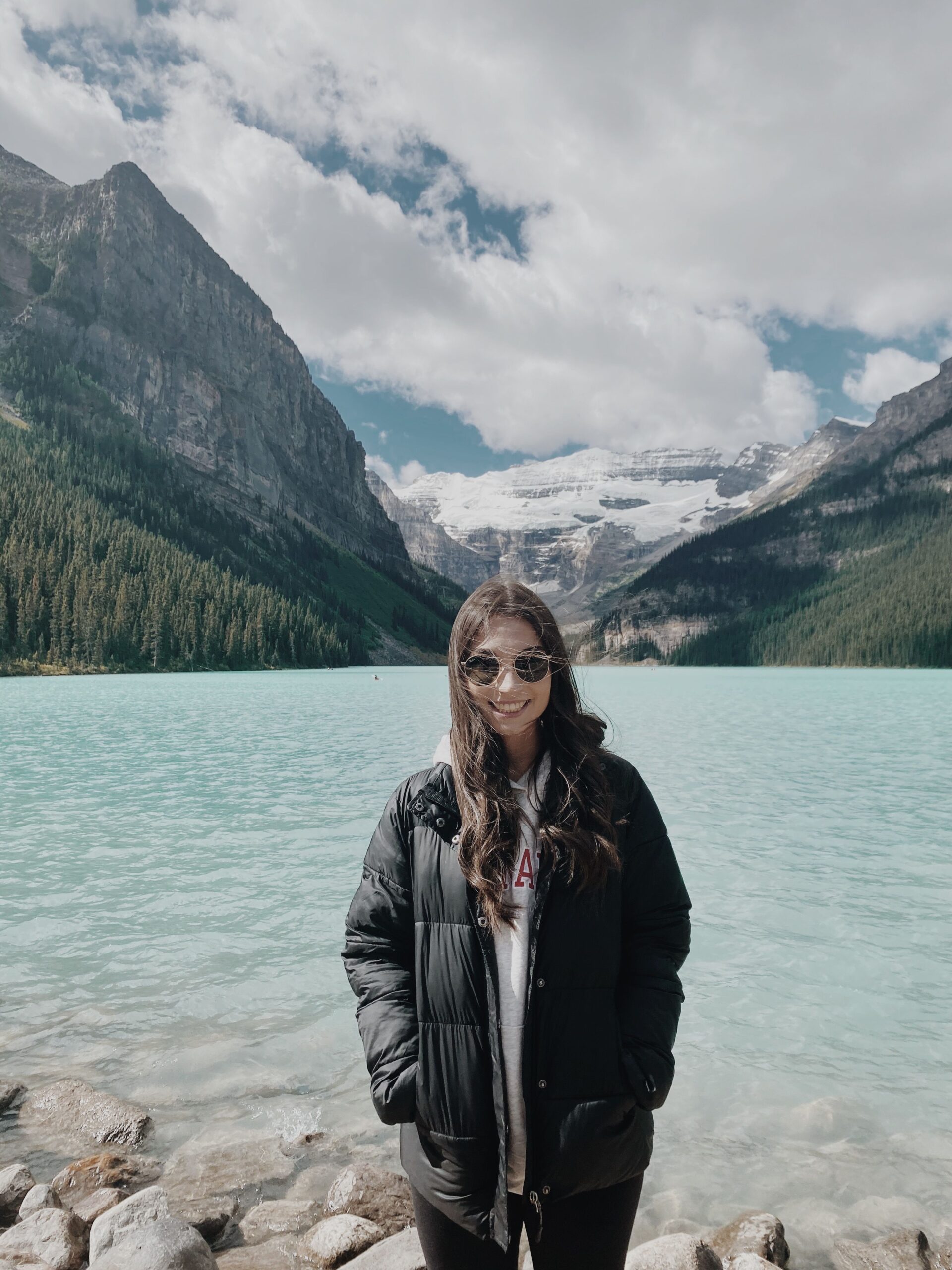 A casual photo of Danielle Tataryn standing in front of Lake Louise, Alberta, a blue green body of water with snowy mountains in the background