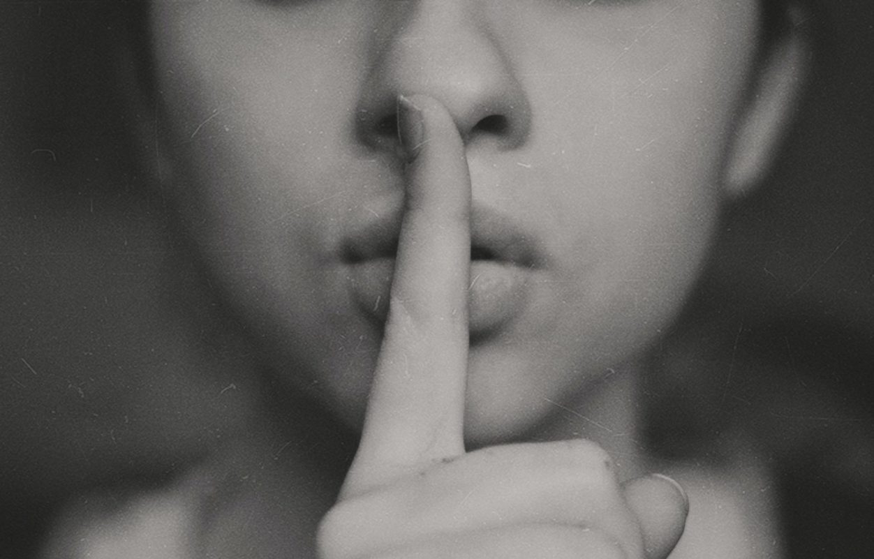 a woman holding her index finger over her lips to indicate a secret that needs to stay quiet, in greyscale