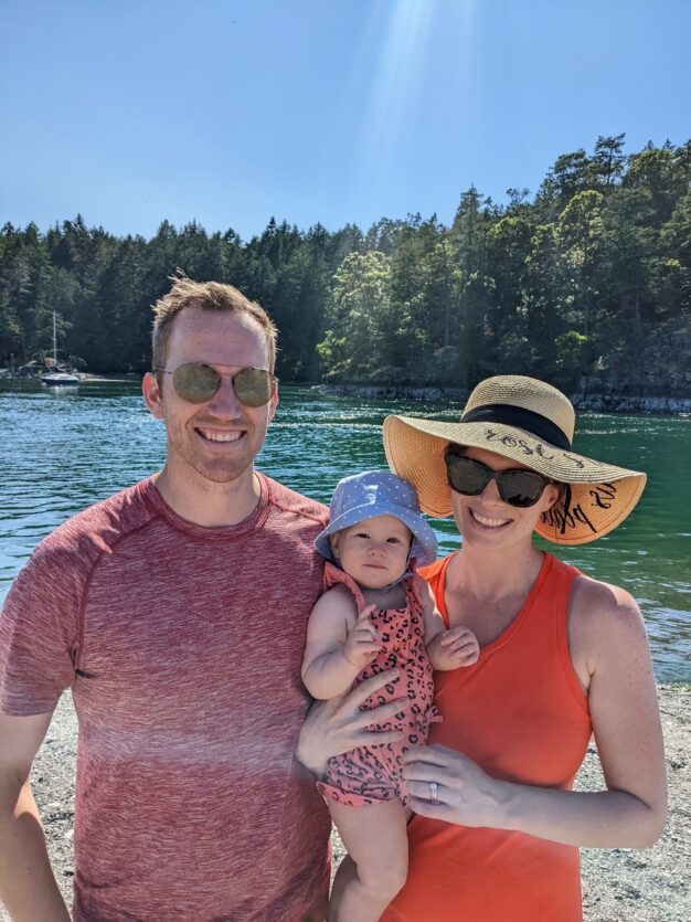 Casual photo of Rosie Jacobs, a brunette in a straw sun hat and orange tank top, holding her baby in a blue hat and pink leopard print onesie, standing next to her husband at the beach.