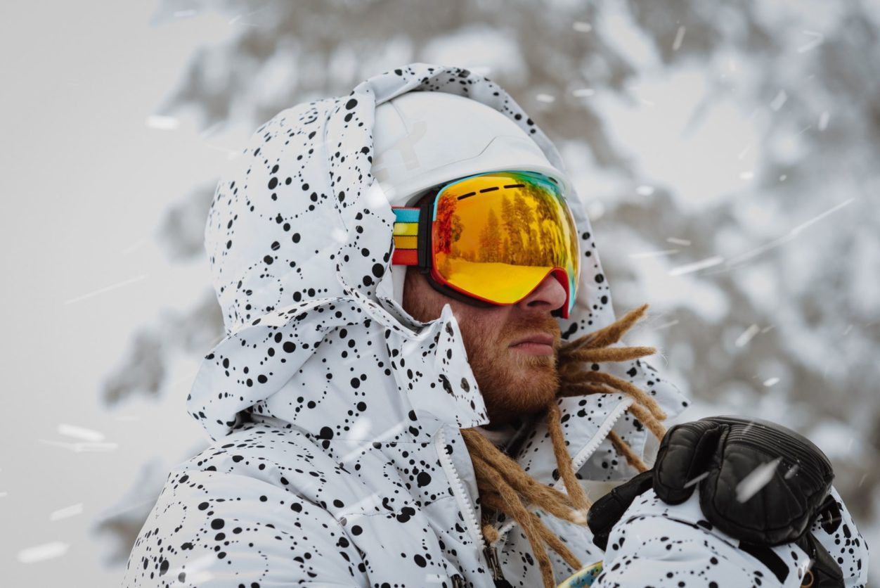 a young red haired man with dreadlocks and yellow ski goggles, and a black and white spotted ski jacket, wearing a helmet