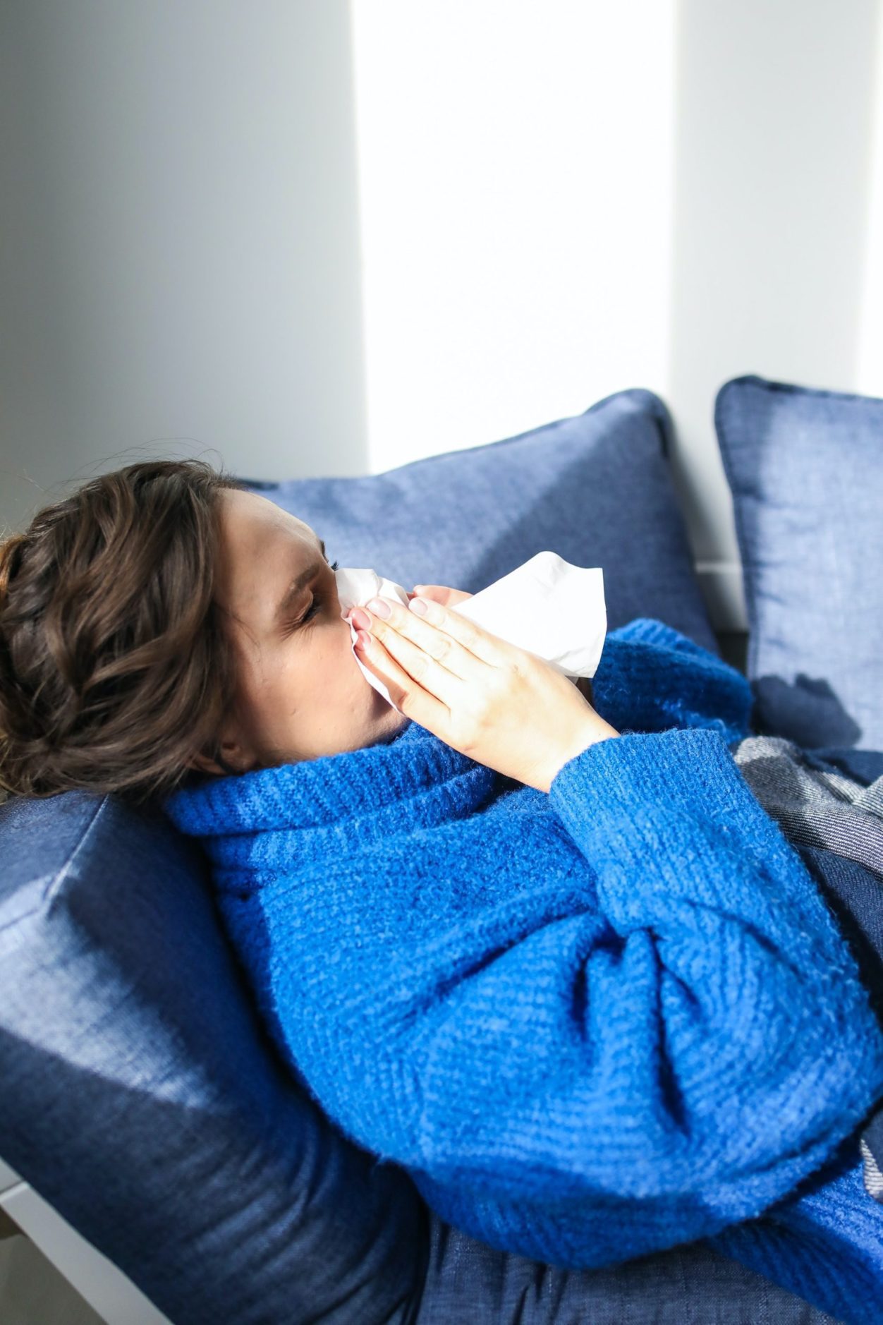 Woman wearing a blue sweater, laying on a sofa and blowing her nose into a tissue relating to paid sick leave for BC employees.