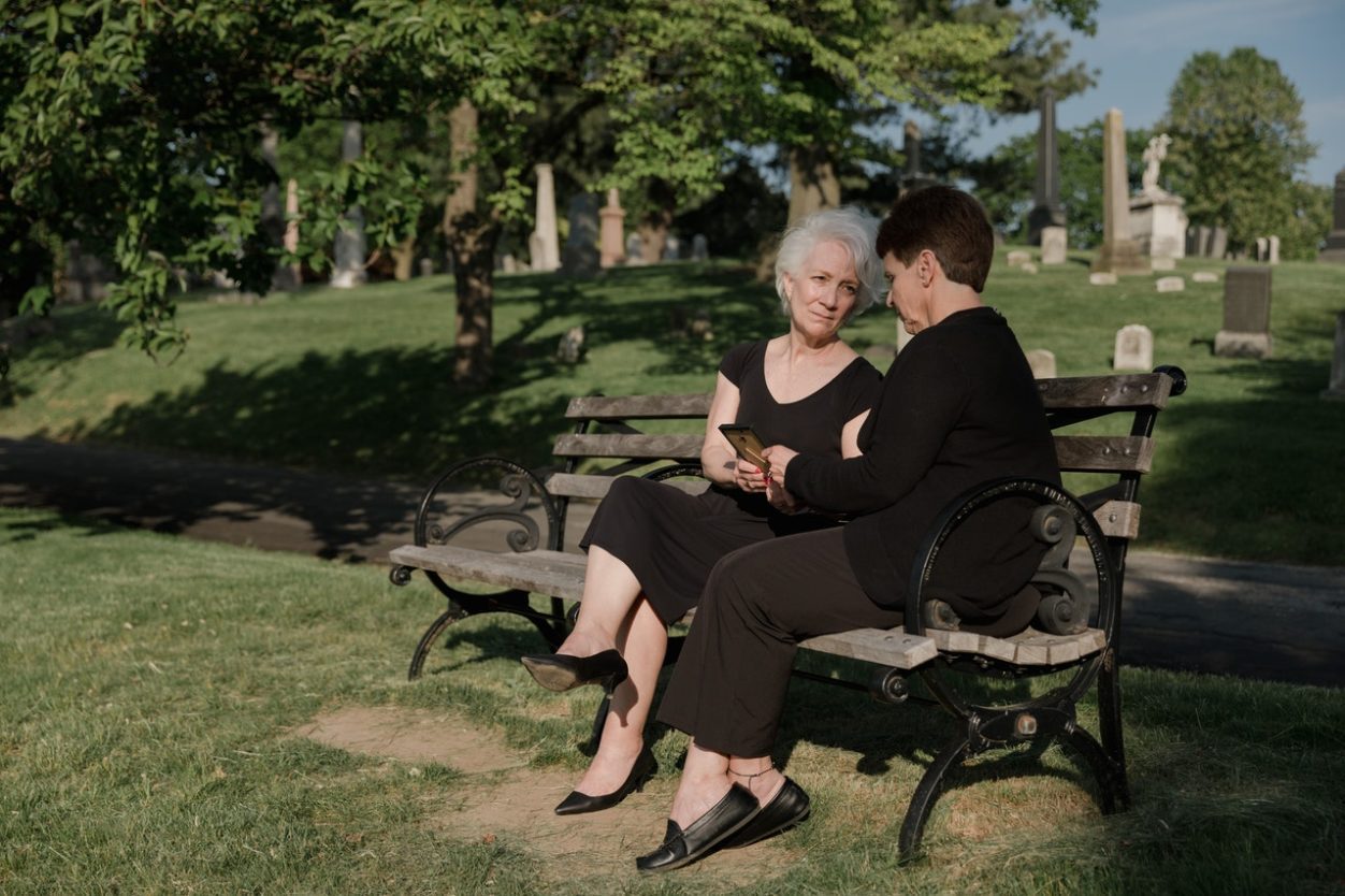 Two women sitting on a wooden bench near a cemetery.